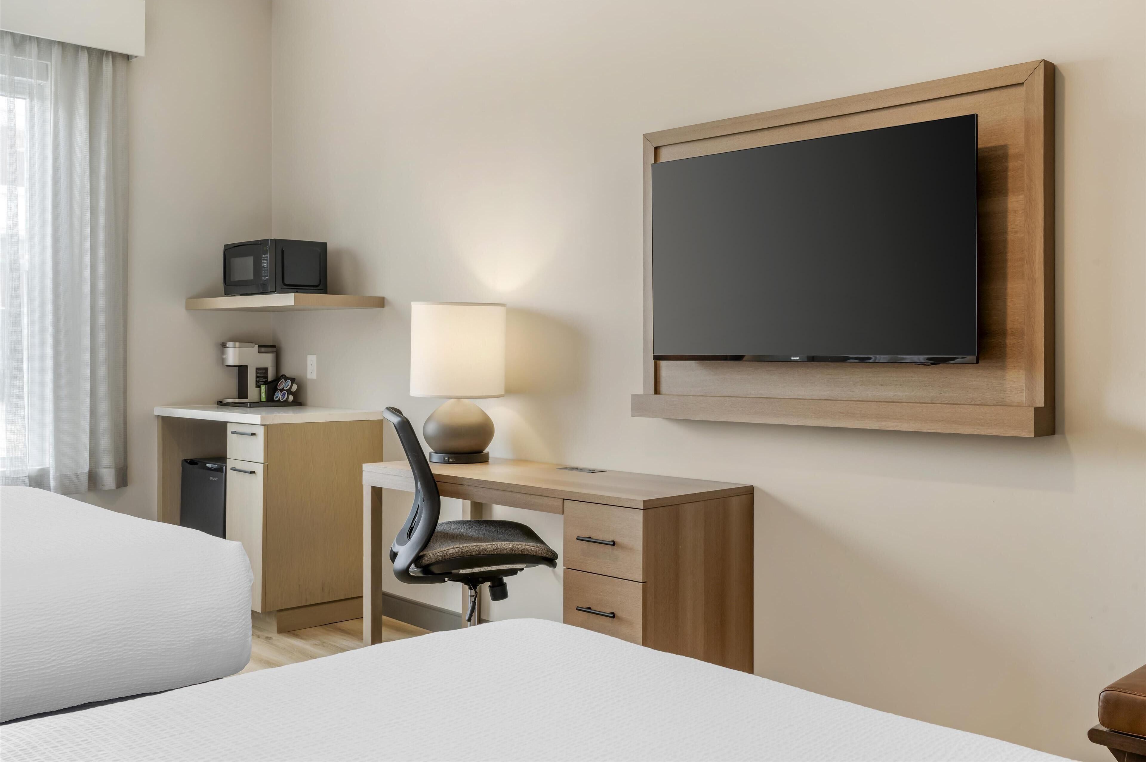 A hotel room with a bed, a desk with a chair, a television mounted on the wall, a lamp, and a coffee machine.