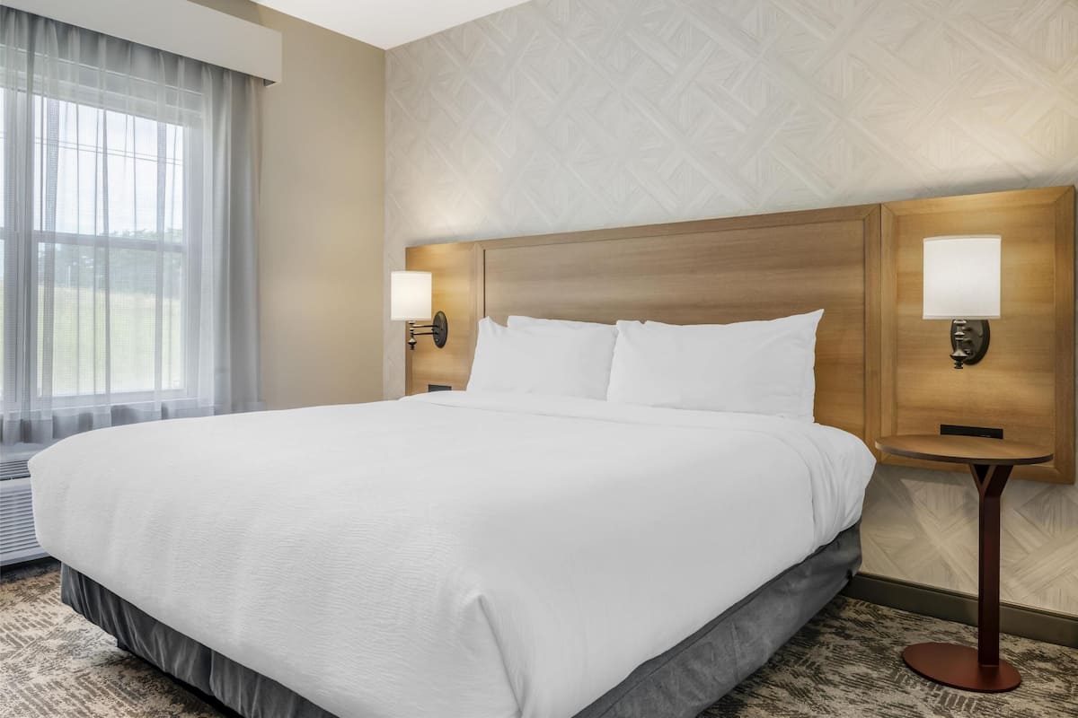 A large bed with white linens, two bedside lamps, and a window with sheer curtains.
