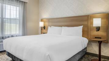 A large bed with white linens, two bedside lamps, and a window with sheer curtains.