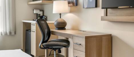 A hotel room with a bed, a desk with a lamp, a chair, a television, a coffee machine, and artwork on the wall.