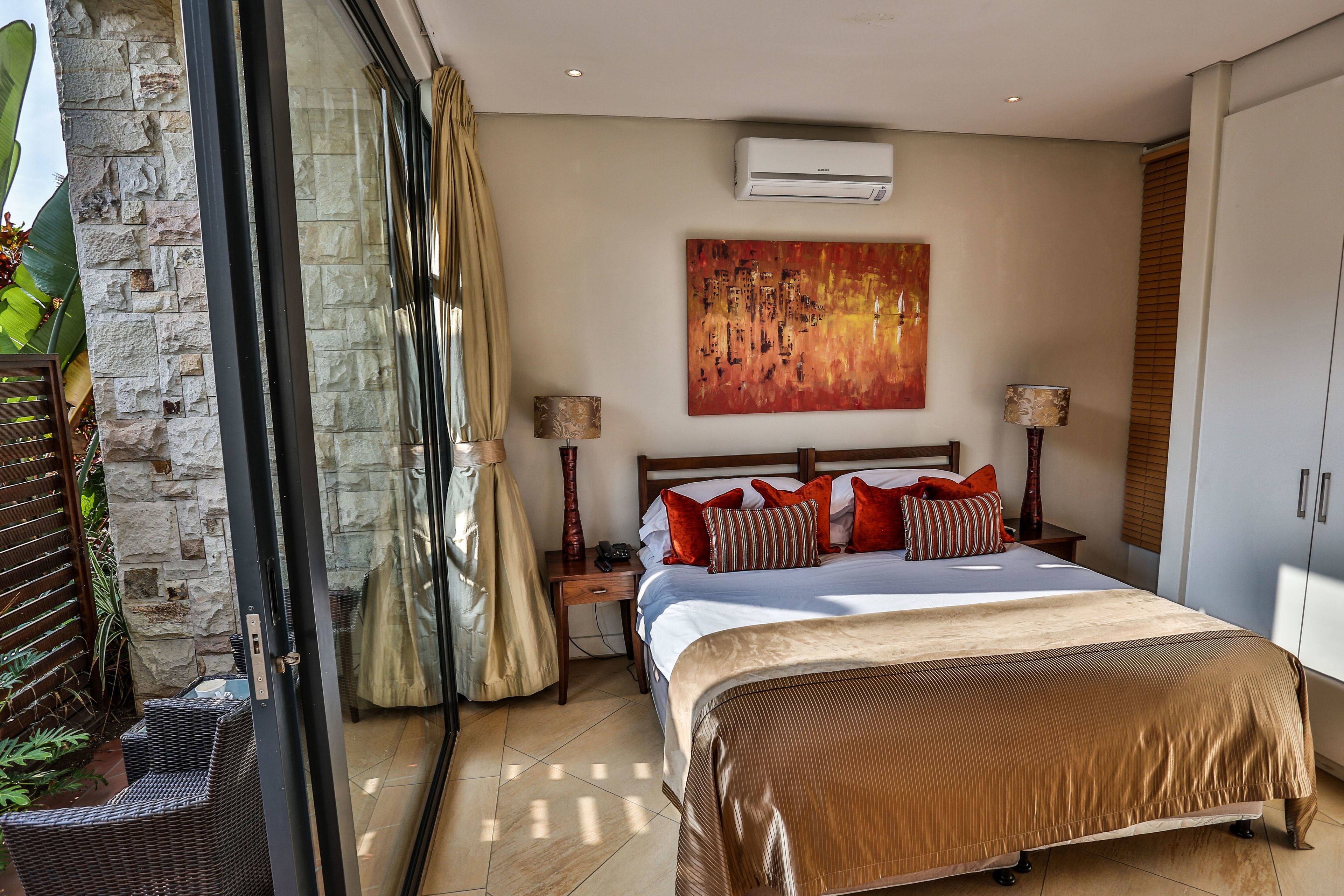 A bedroom with a large bed, a nightstand, a lamp, a painting, and a sliding glass door leading to a balcony with plants.