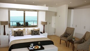 A modern bedroom with a large bed, two chairs, a small table, and a tray with food and drink. A window offers a view of the ocean. - Canelands Beach Club (Ballito)