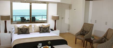 A modern bedroom with a large bed, two chairs, a small table, and a tray with food and drink. A window offers a view of the ocean.