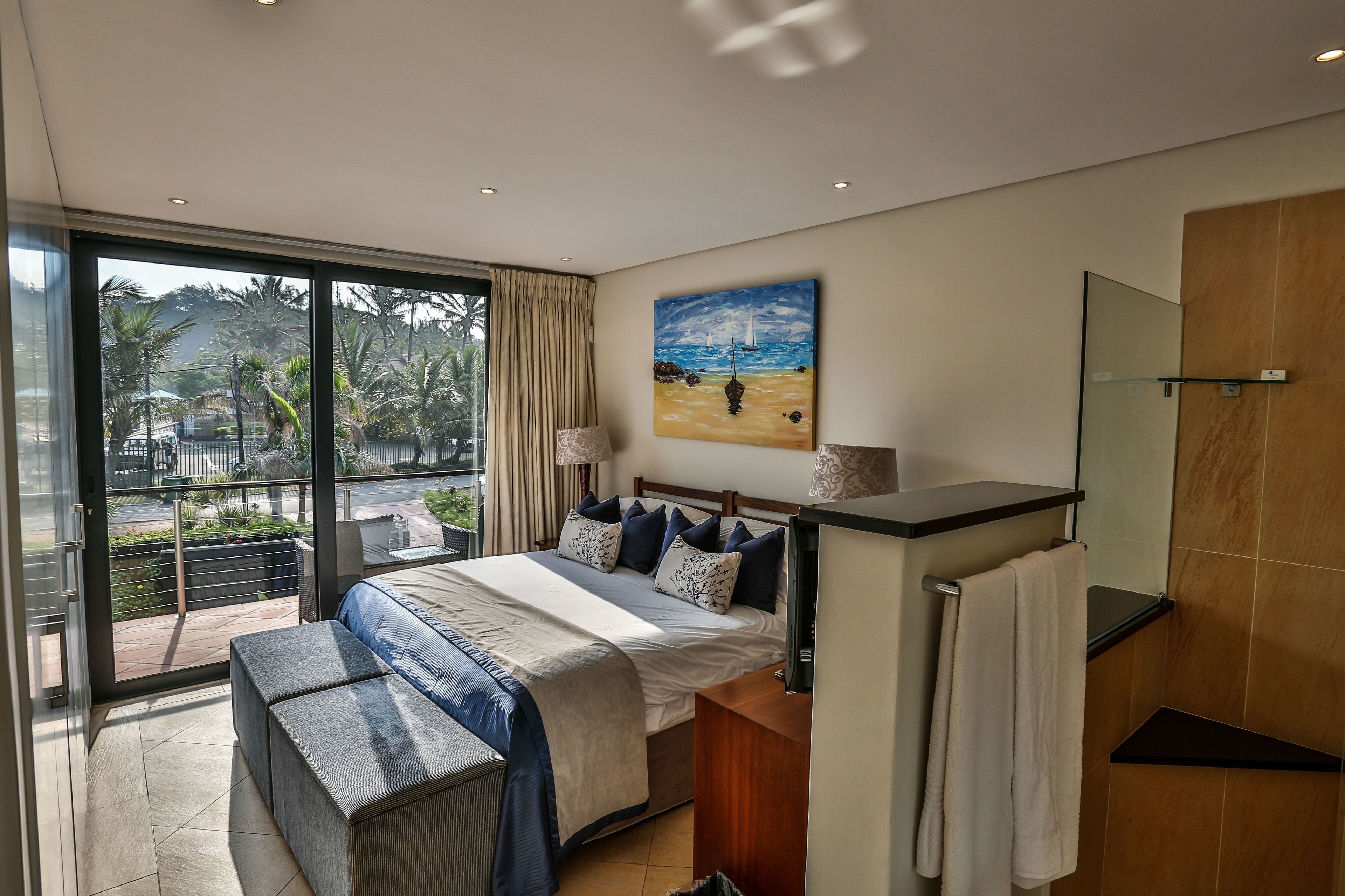 A modern bedroom with a large bed, a bench, a bathroom with a mirror, and a balcony with a view of palm trees and a beach.