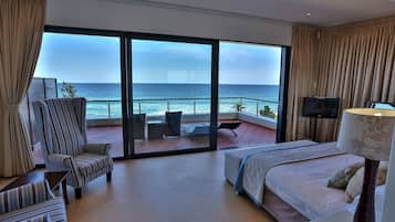A hotel room with a large sliding glass door leading to a balcony with a view of the ocean. There is a bed, a chair, a TV, and a lamp on a bedside table.
