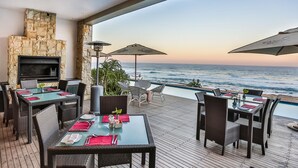 A deck with tables and chairs, a stone fireplace, and a view of the ocean. - Canelands Beach Club (Ballito)