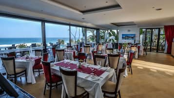 Un restaurante con mesas dispuestas para comer, amplios ventanales que ofrecen vista al océano y una cortina roja a la derecha.