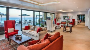 A spacious living area with a large glass wall overlooking the sea, furnished with red and beige sofas, a coffee table, and a fireplace. - Canelands Beach Club (Ballito)