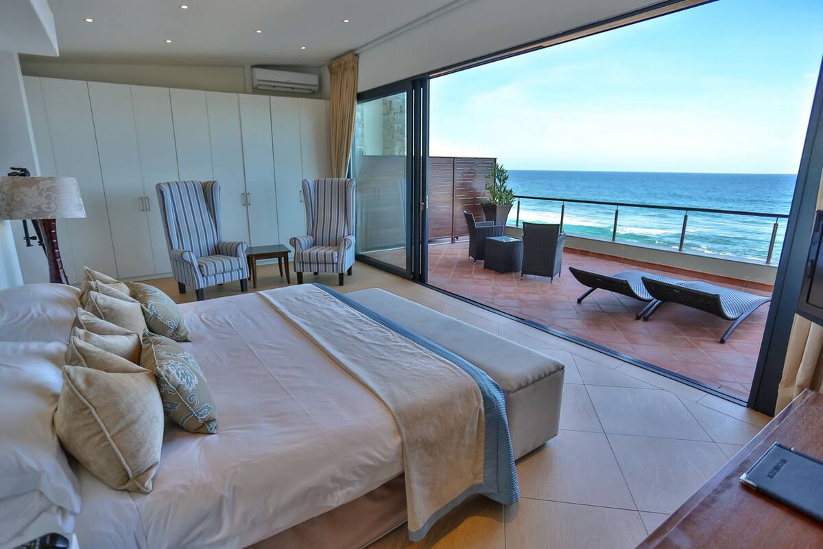 Ein Schlafzimmer mit einem großen Bett, zwei Sesseln, einem kleinen Tisch und Meerblick durch eine Schiebetür.