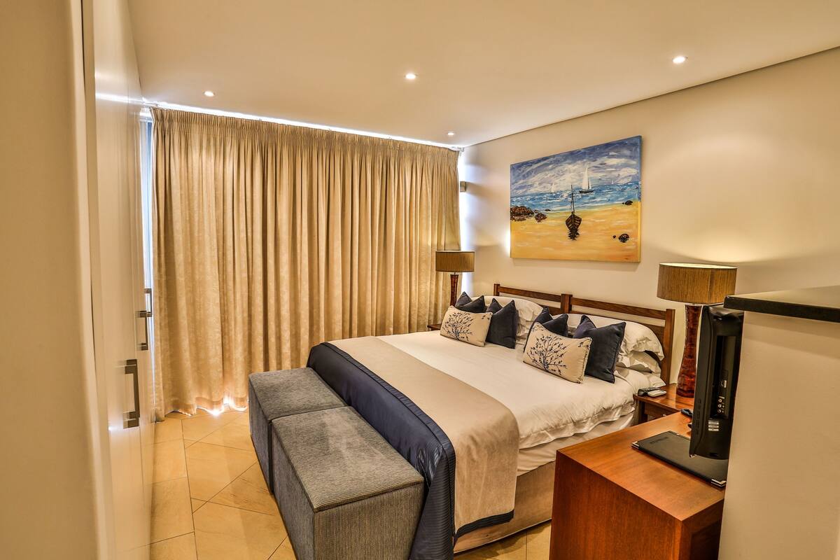 Ein Hotelzimmer mit einem großen Bett, einem Nachttisch mit einem Laptop, einer Bank und einem Strandbild.