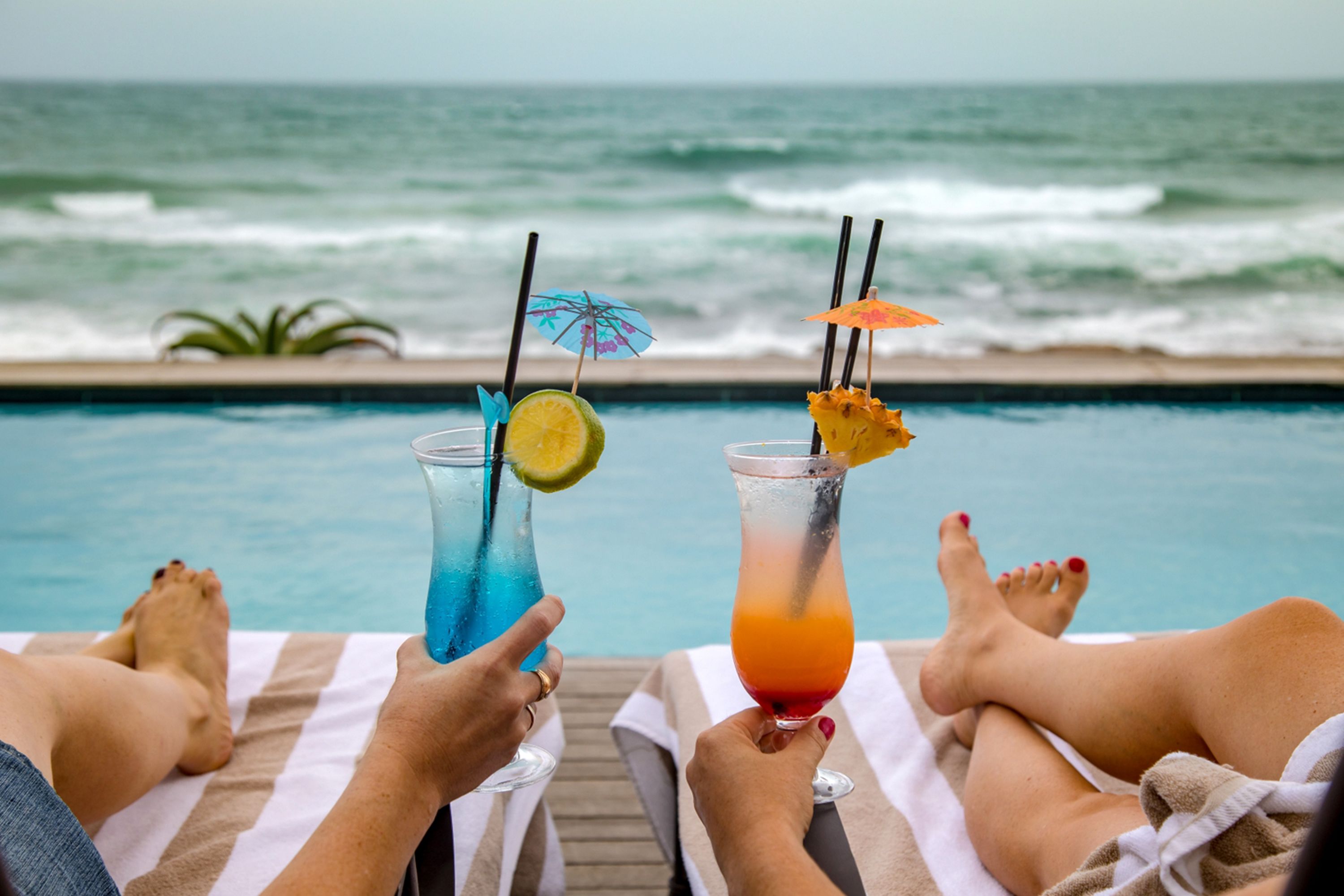 Dos personas descansando junto a una piscina con bebidas, con vista al océano.