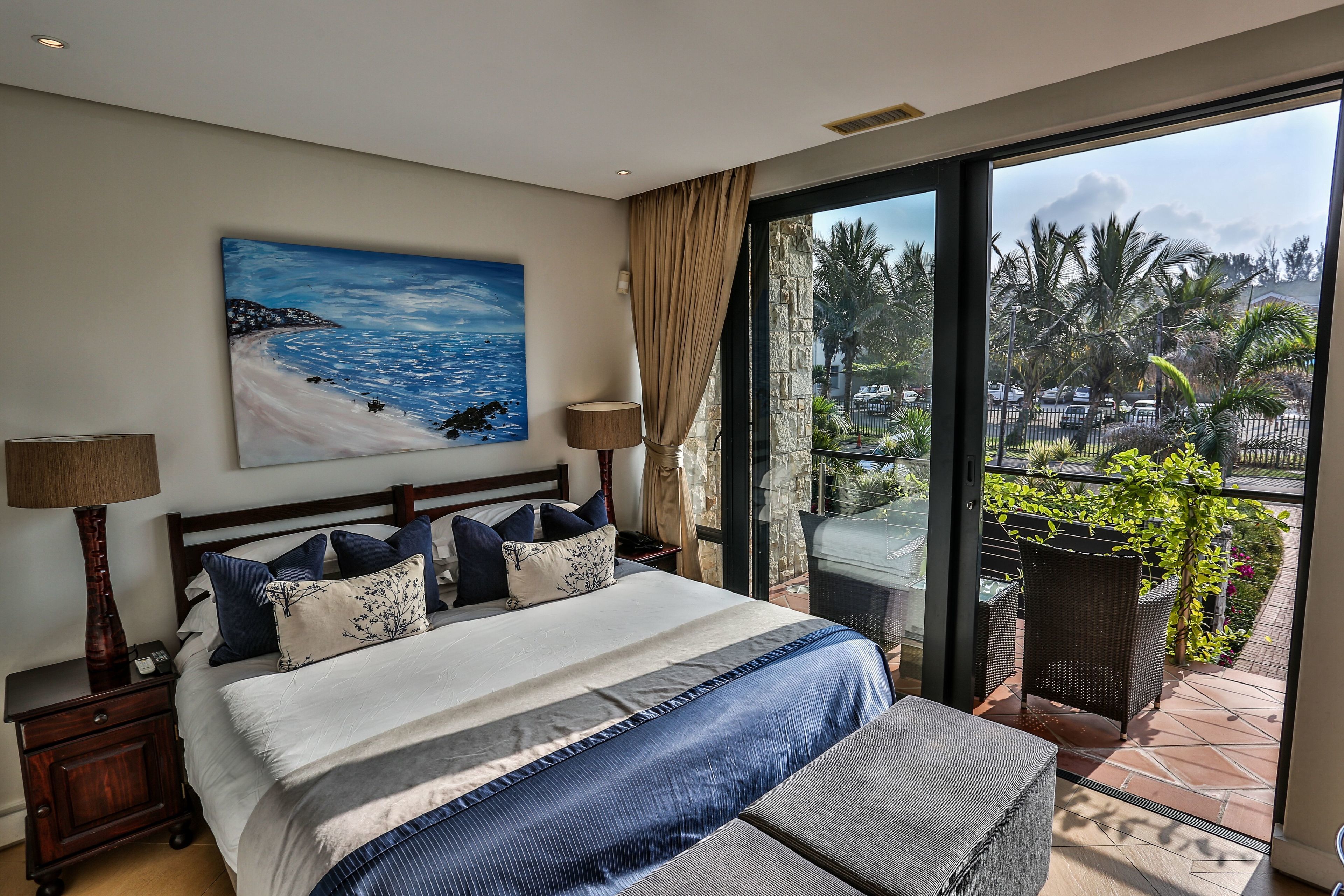 A large bed with a view of the outdoors, a painting on the wall, and a balcony with plants.
