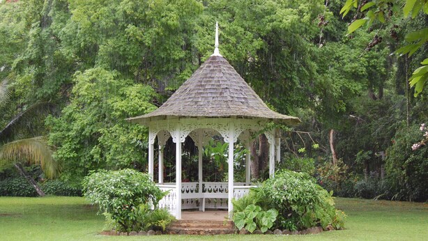 Um gazebo com telhado em azulejos e grades brancas, rodeado por vegetação exuberante e árvores.