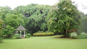 Um gazebo num jardim exuberante, com árvores densas e grama bem cuidada.