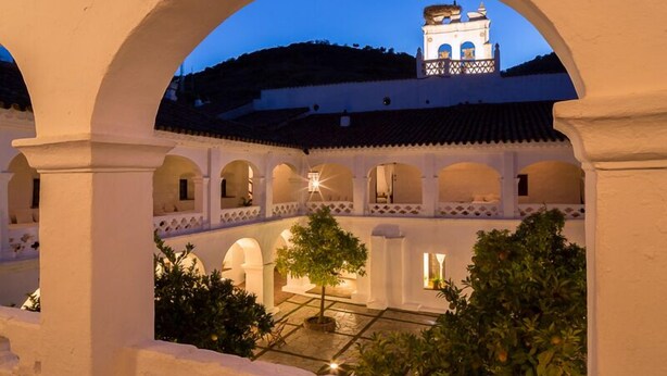 Un patio arquitectónico con arcos, un árbol en el centro y una torre de campanas a lo lejos.