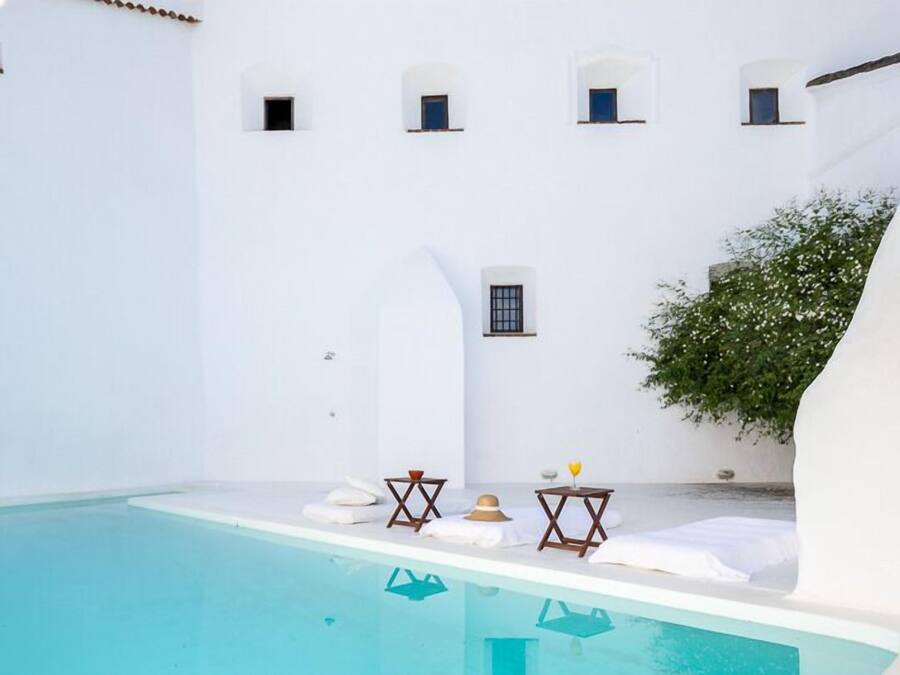 Un espace piscine aux murs blancs, avec une piscine à l’eau d’un bleu limpide, de petites tables avec des parasols et une plante en pot.