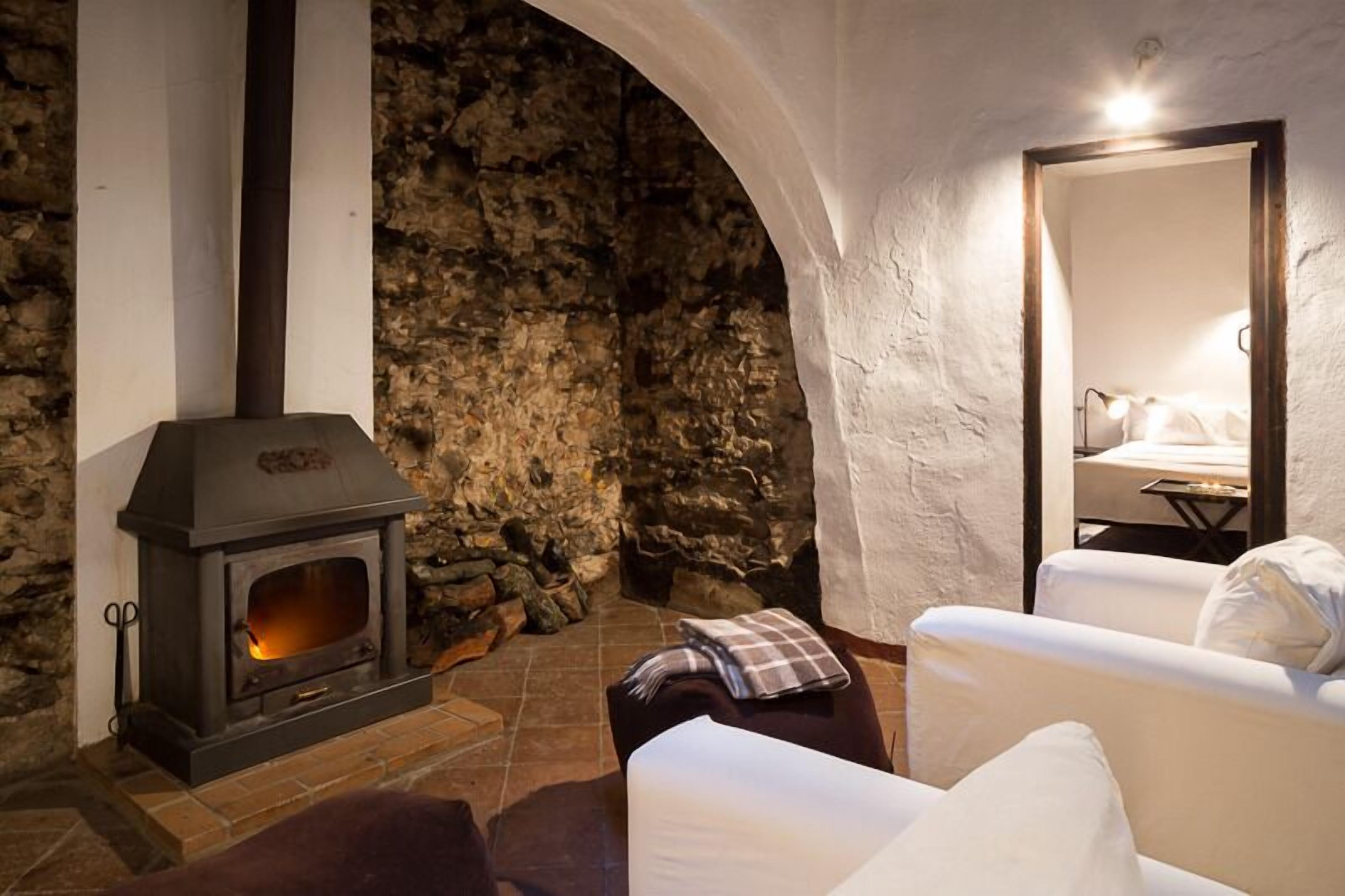 Habitación de paredes de piedra con chimenea, un sillón blanco y una cama con dosel blanco.