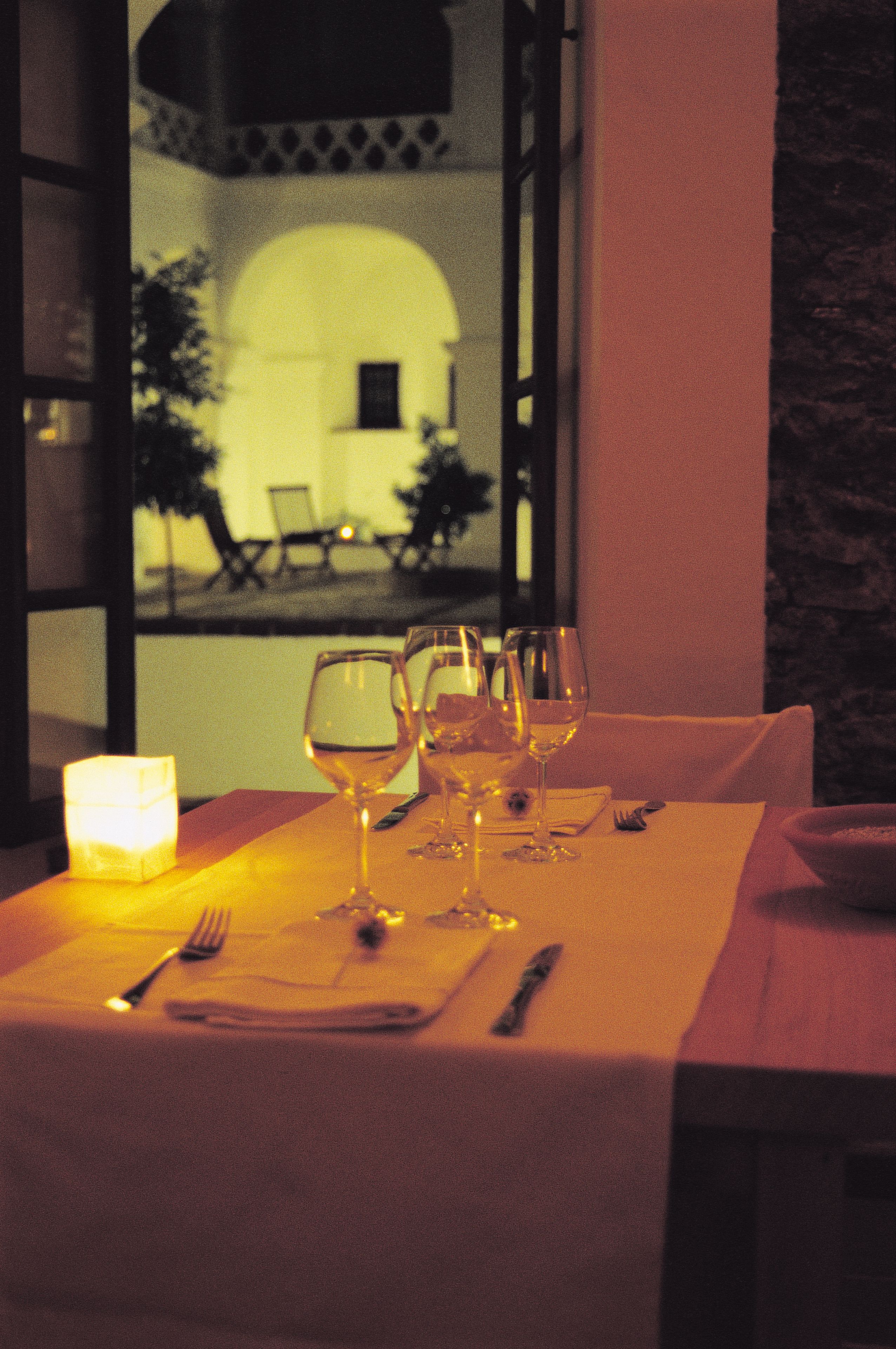 Mesa posta para refeição com copos de vinho e uma vela, com vista para um pátio com cadeiras e um edifício.