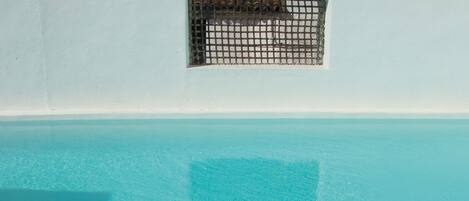 Una piscina con agua azul cristalina, un reposapiés de mimbre con una pequeña maceta y toallas blancas en sillas de descanso.