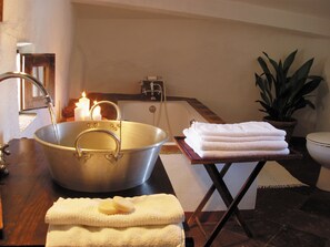 Un baño con un lavamanos metálico grande, una pila de toallas blancas, una vela encendida y una planta en maceta.