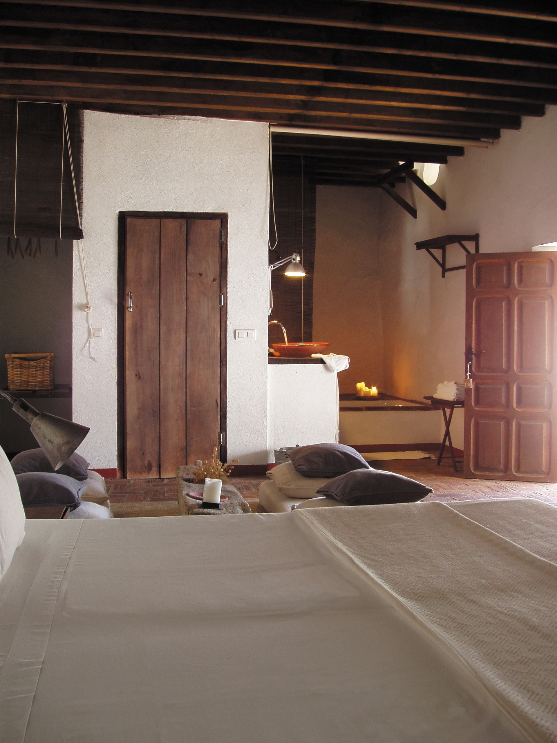 Una habitación rústica con una puerta de madera, una mesita con una vela y una cama con ropa de cama blanca.