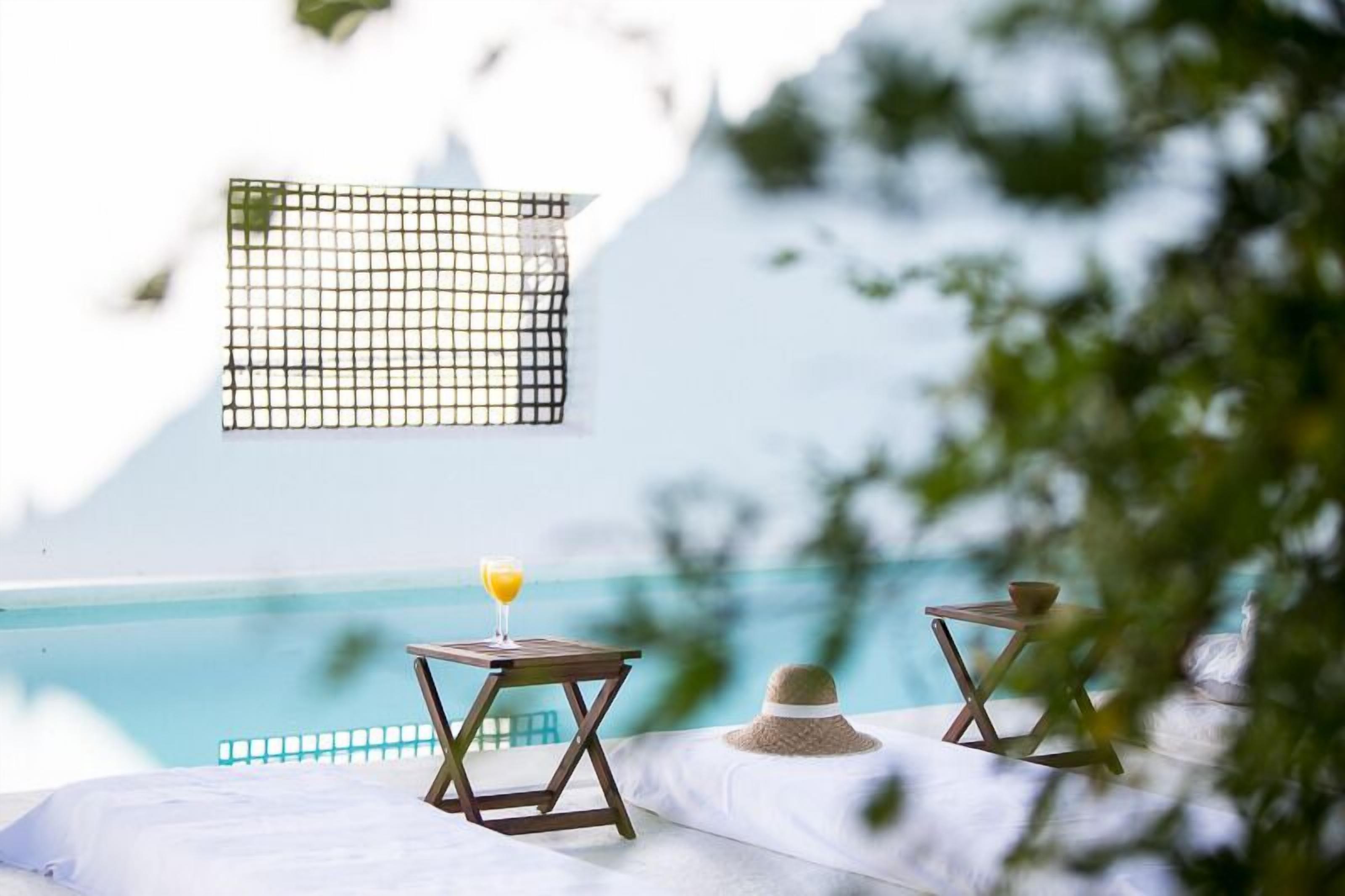 A poolside area with a view of the sea, two wooden tables with drinks, and a straw hat on a cushion.