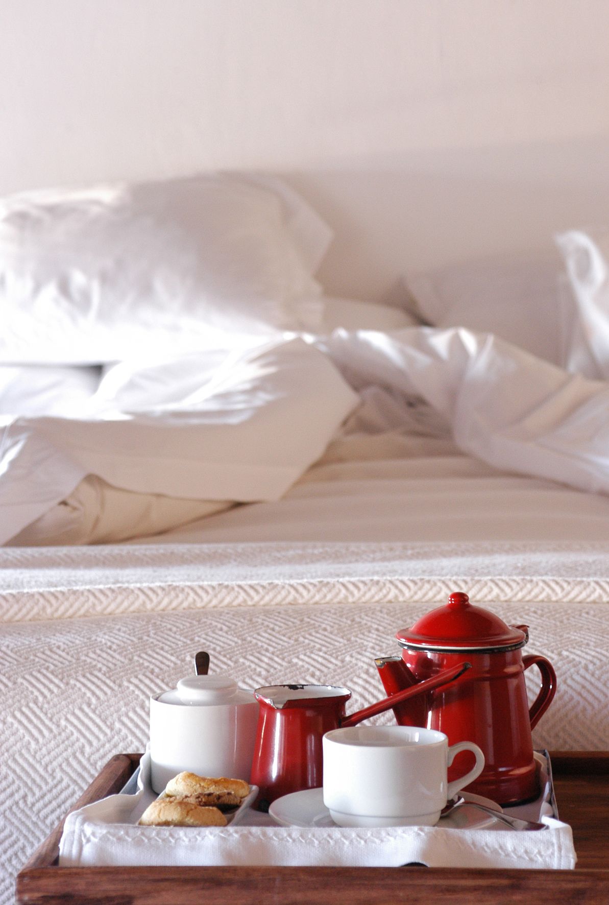 A breakfast tray with a red coffee pot, white cups, and biscuits on a wooden tray, placed on a bed with white bedding.