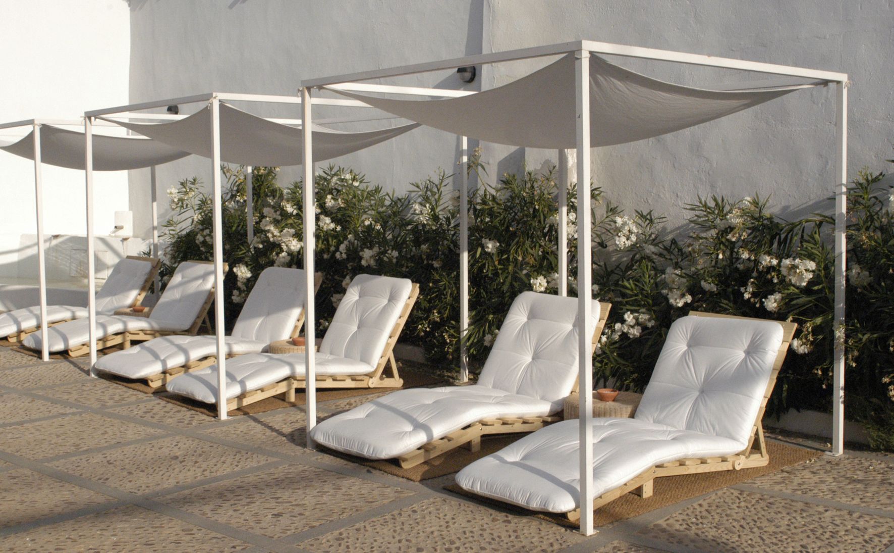 A row of sun loungers with white cushions and wooden frames, shaded by white canopies, against a backdrop of white flowers and a plain wall.