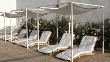 A row of sun loungers with white cushions and wooden frames, shaded by white canopies, against a backdrop of white flowers and a plain wall.