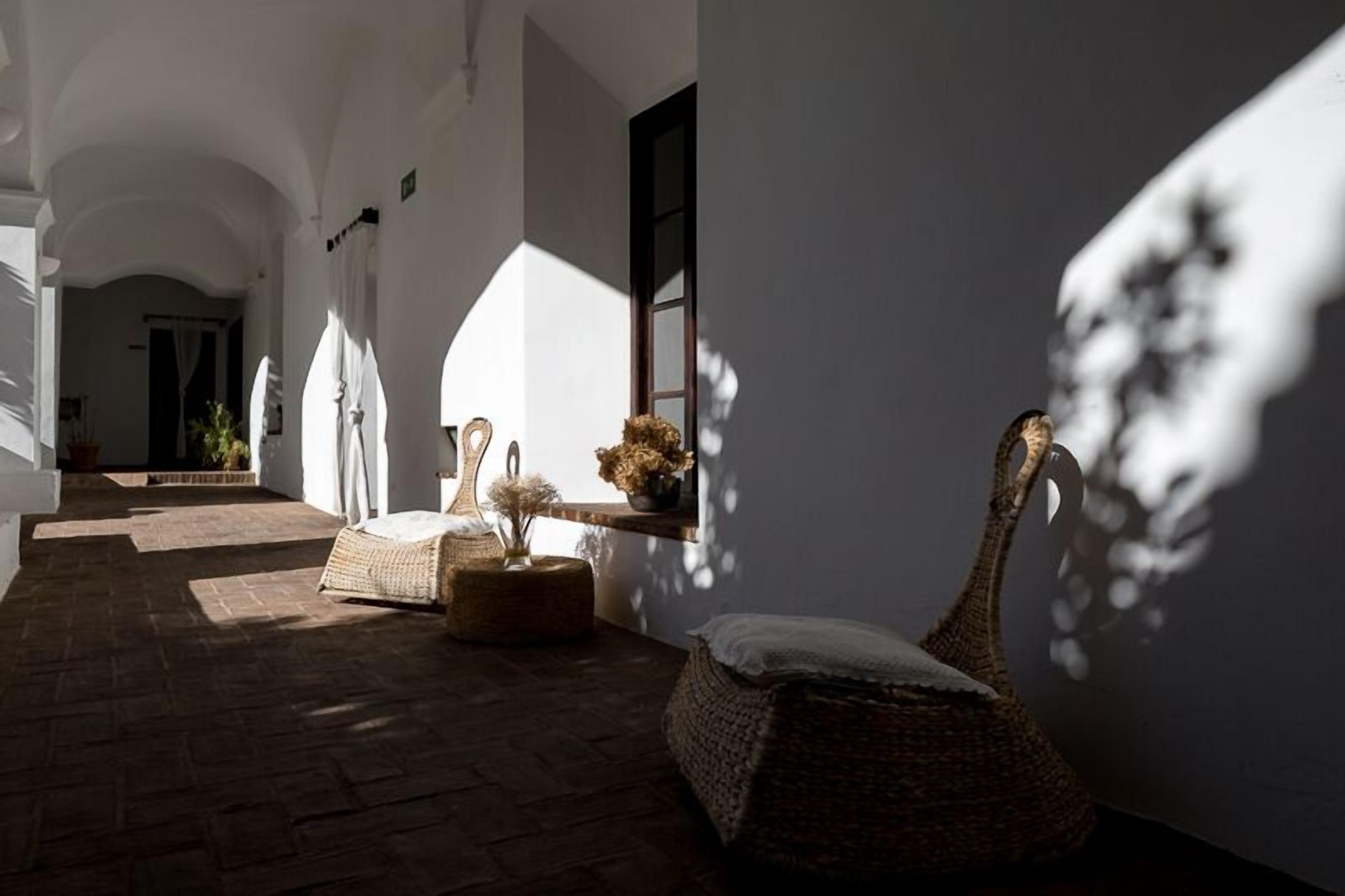 Un pasillo iluminado por el sol con muebles de mimbre, una pequeña mesa con flores y una ventana que proyecta sombras.