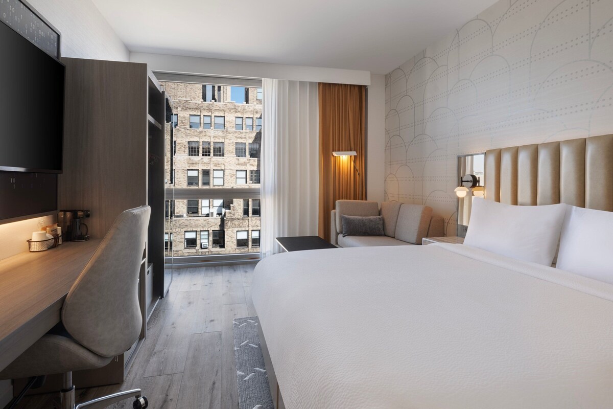 Une chambre d’hôtel comprenant un lit, un bureau, une chaise, un téléviseur, une fenêtre donnant sur des immeubles et un mur orné d’un motif géométrique.