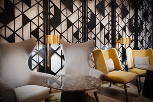 Egy modern, elegáns pihenősarok, geometriai mintázatú falakkal, egy kerek márványasztallal és két, lámpákkal felszerelt karosszékkel.