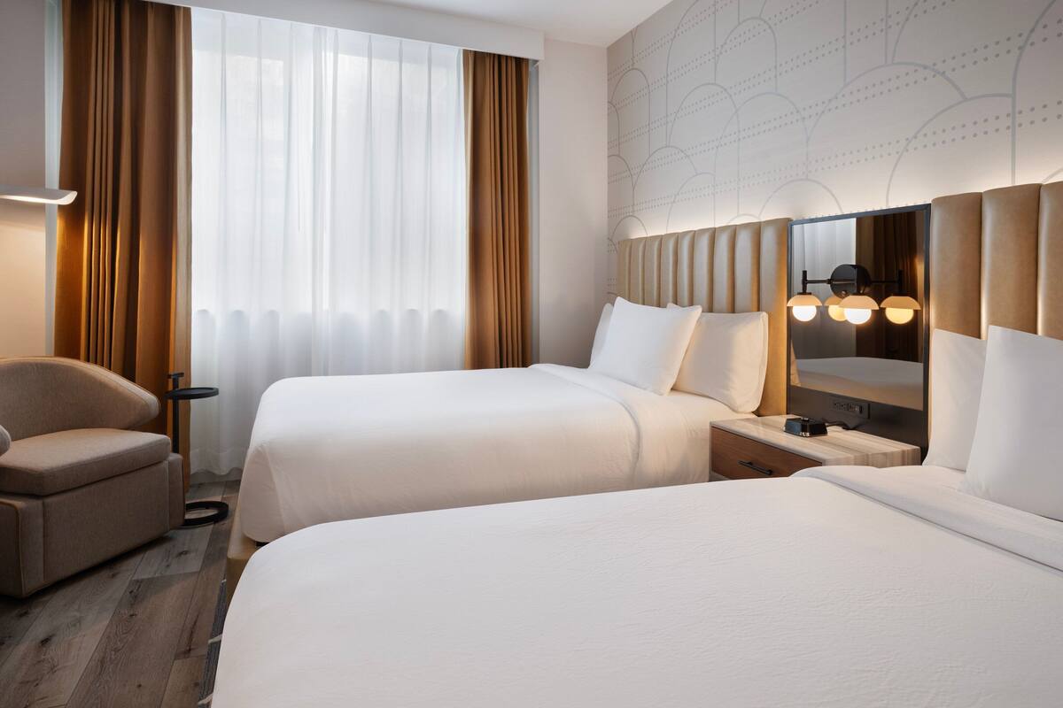 A hotel room with two beds, a chair, a nightstand, a mirror, and a window with curtains.