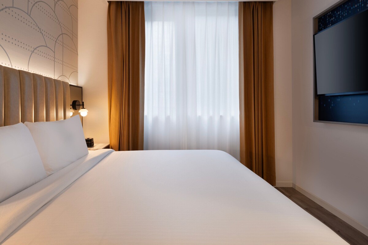 Een hotelkamer met een groot bed, een hoofdeinde met een lamp, een nachtkastje, een aan de muur bevestigde televisie en een raam met dunne gordijnen.