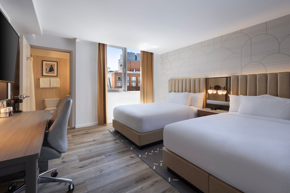Une chambre d’hôtel avec deux lits, un bureau, une chaise, une télévision et une fenêtre donnant sur la ville.