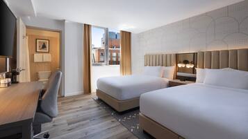 A hotel room with two beds, a desk, a chair, a television, and a window with a city view.