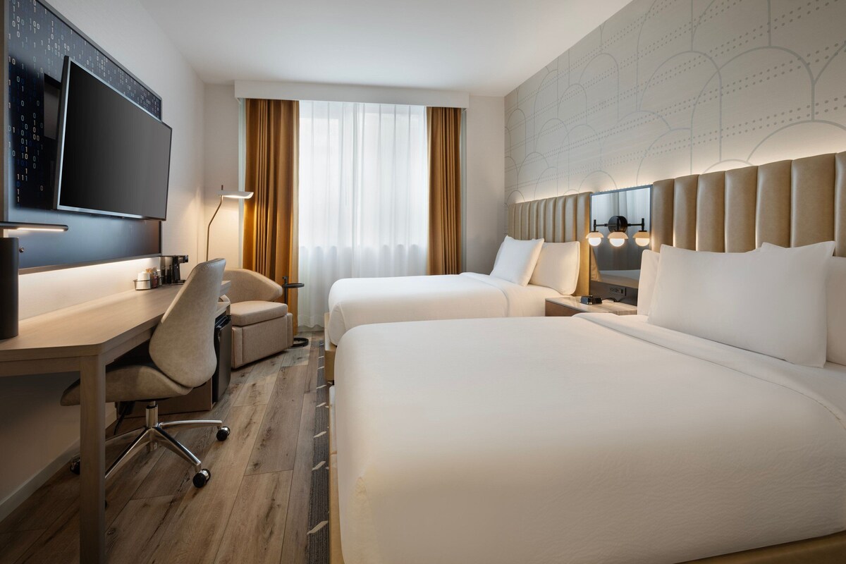 Une chambre d’hôtel avec deux lits, un bureau, une chaise, une télévision et une grande fenêtre avec des rideaux.