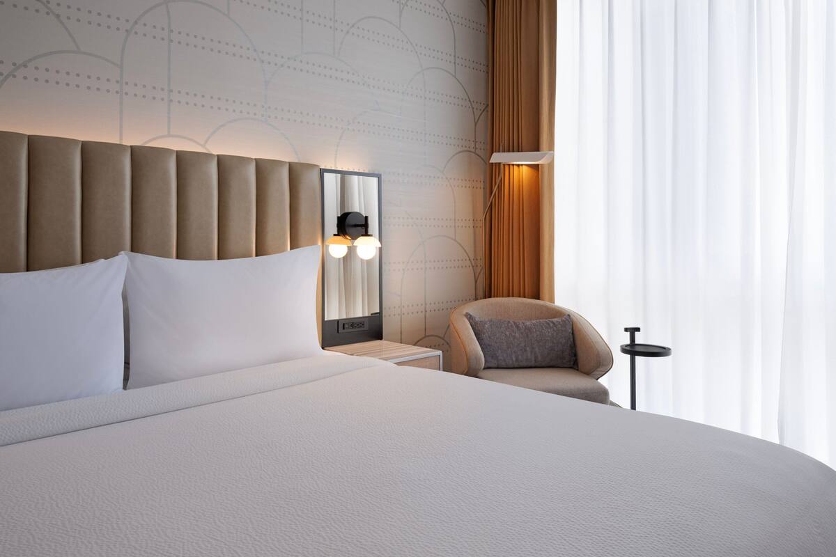 A modern hotel room with a large bed, a headboard with integrated lighting, a side table, a chair, and a mirror.