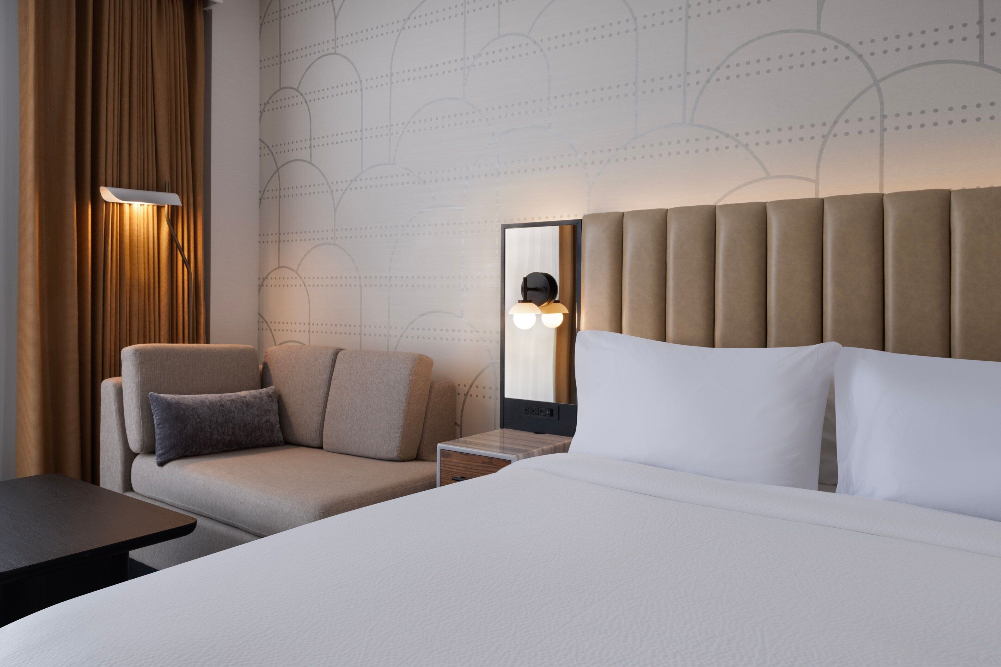 A hotel room with a large bed, a sofa, a small table, and a wall-mounted mirror.