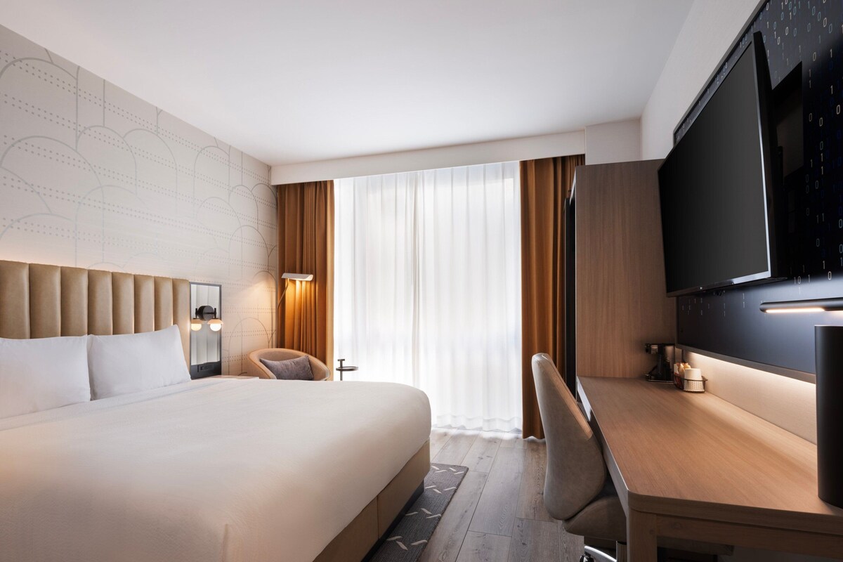 Une chambre d’hôtel moderne dotée d’un grand lit, d’un bureau, d’une télévision à écran plat et d’une grande fenêtre avec des rideaux.
