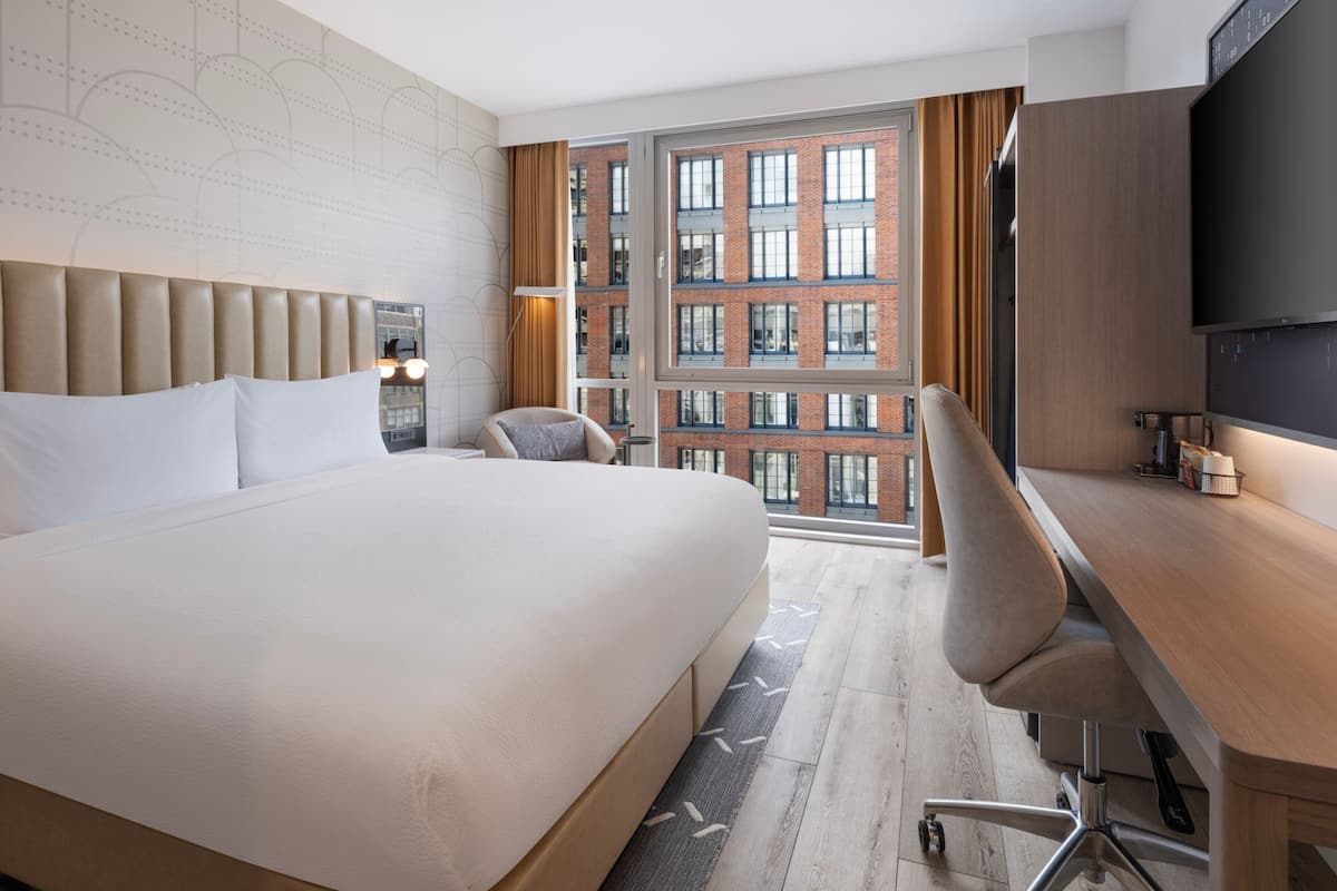 A hotel room with a large bed, a desk, a chair, a TV, and a window with a view of a brick building.
