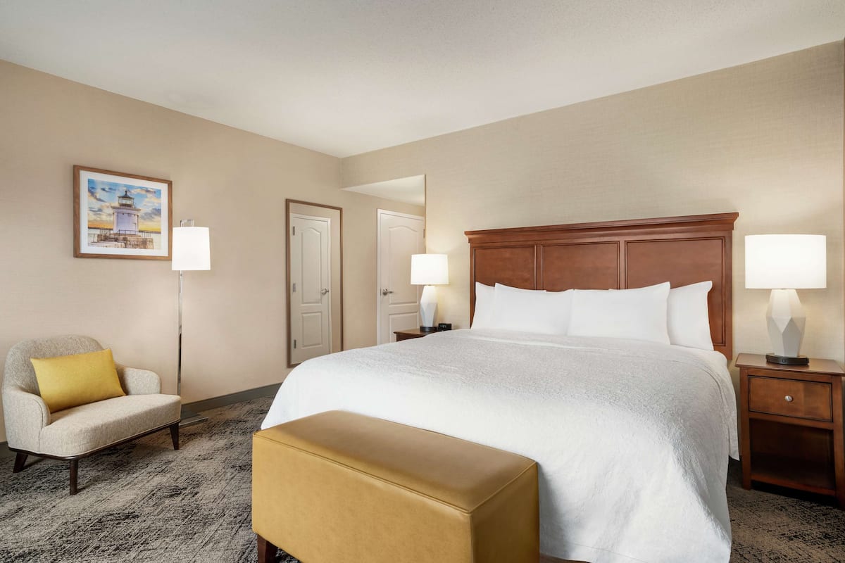 A hotel room with a large bed, a chair, a bench, two bedside tables with lamps, a door, and a framed picture on the wall.