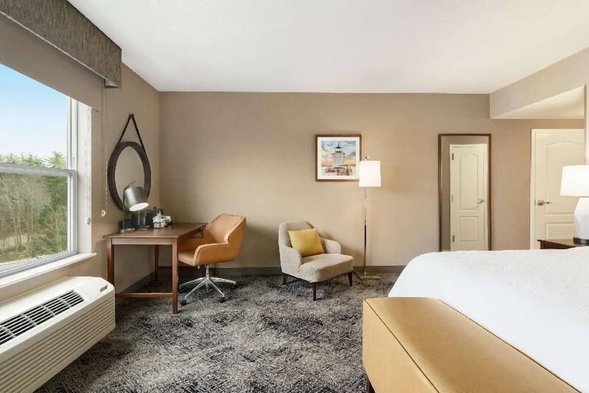 A hotel room with a bed, a desk, a chair, a lamp, a picture on the wall, and a window.