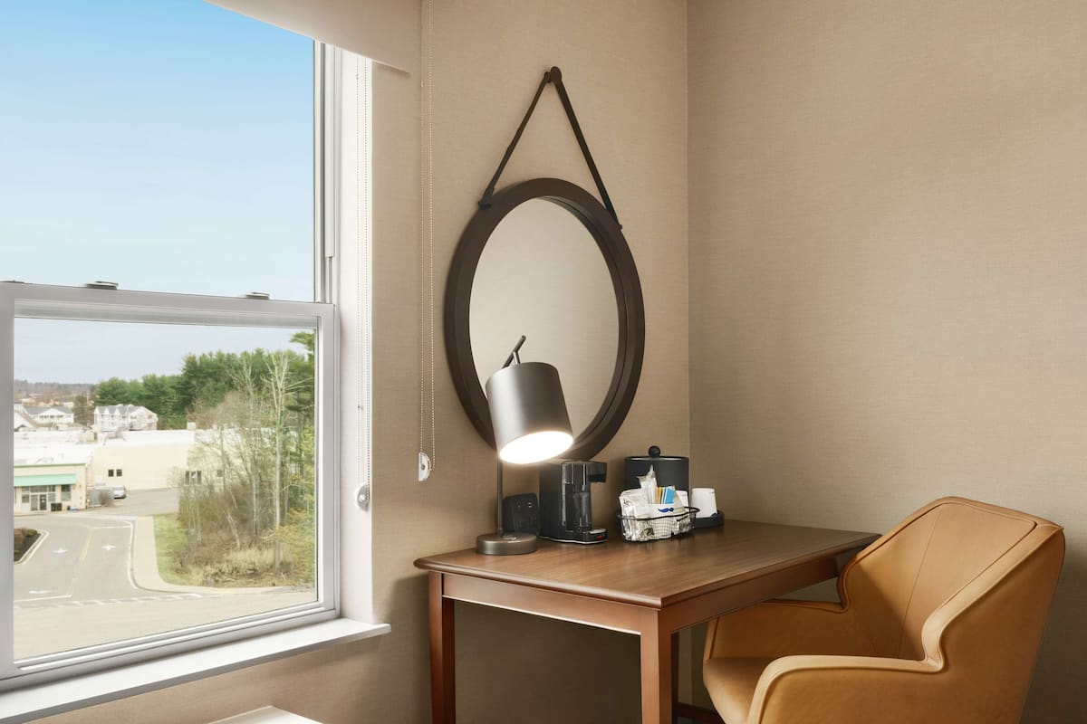 A hotel room with a desk, a chair, a lamp, a coffee maker, and a mirror on the wall.