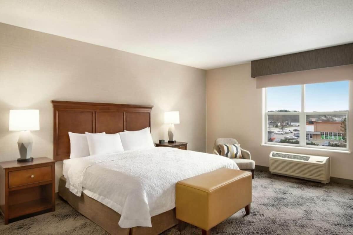 A hotel room with a large bed, two bedside tables with lamps, a chair, a window with a view, and an air conditioning unit.