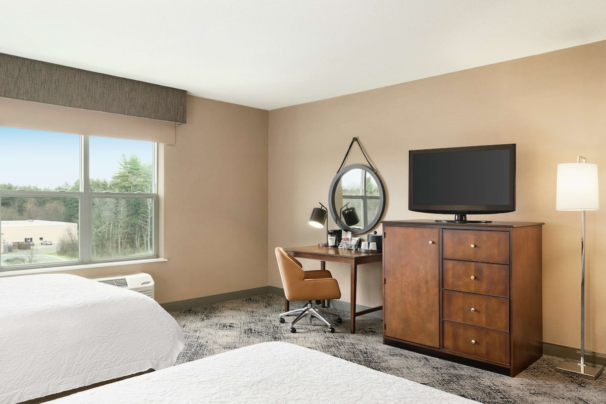 A hotel room with a bed, a desk with a chair, a television, a dresser, and a window with a view of trees.