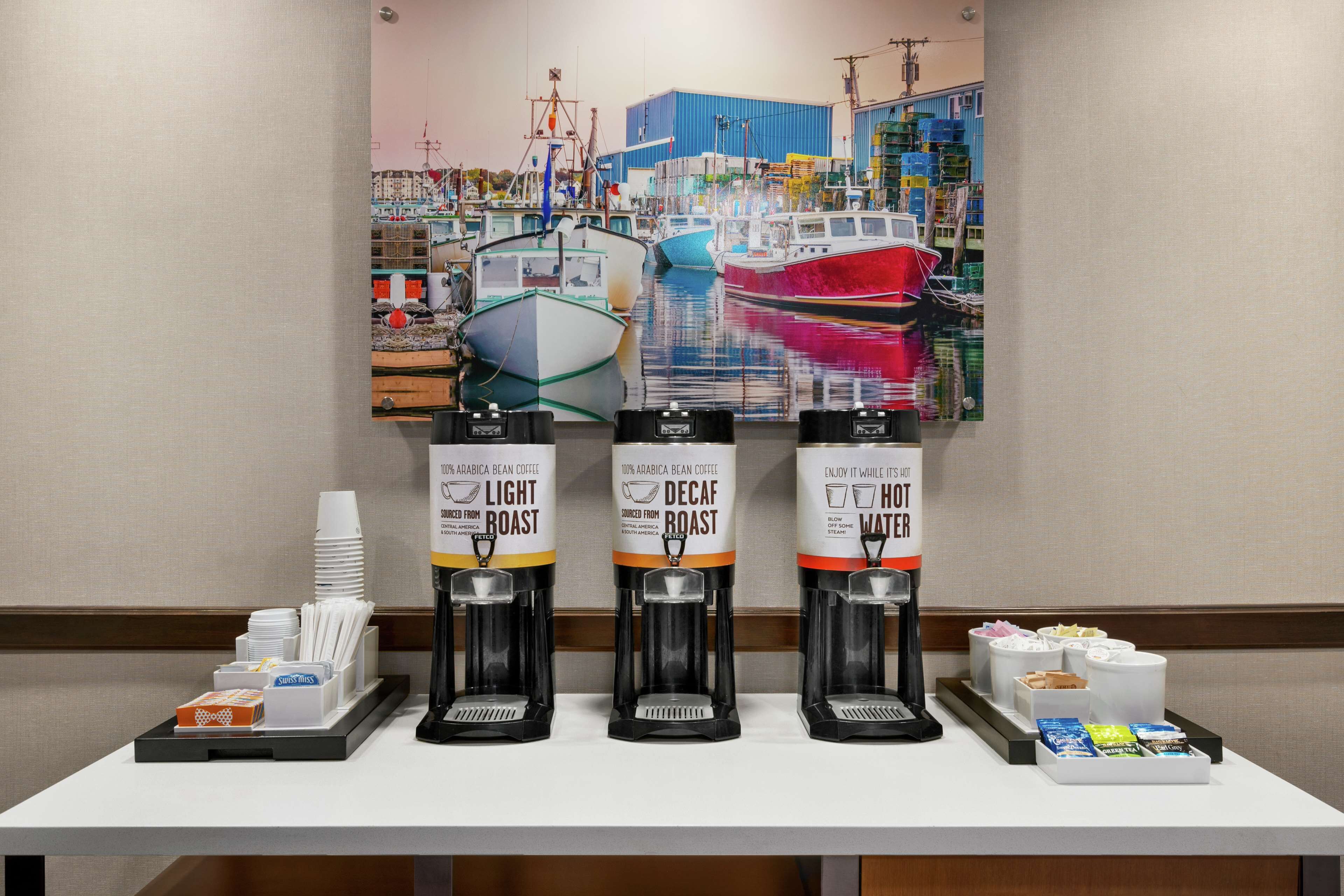 En kaffestation med tre kaffedispensrar märkta med "Ljust rostad", "Koffeinfritt" och "Varmt vatten", samt ett bricka med engångsmuggar och snacks.