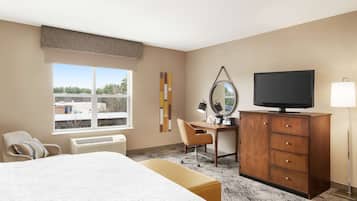 A hotel room with a bed, a desk, a television, a chair, a dresser, and a window with a view of buildings.
