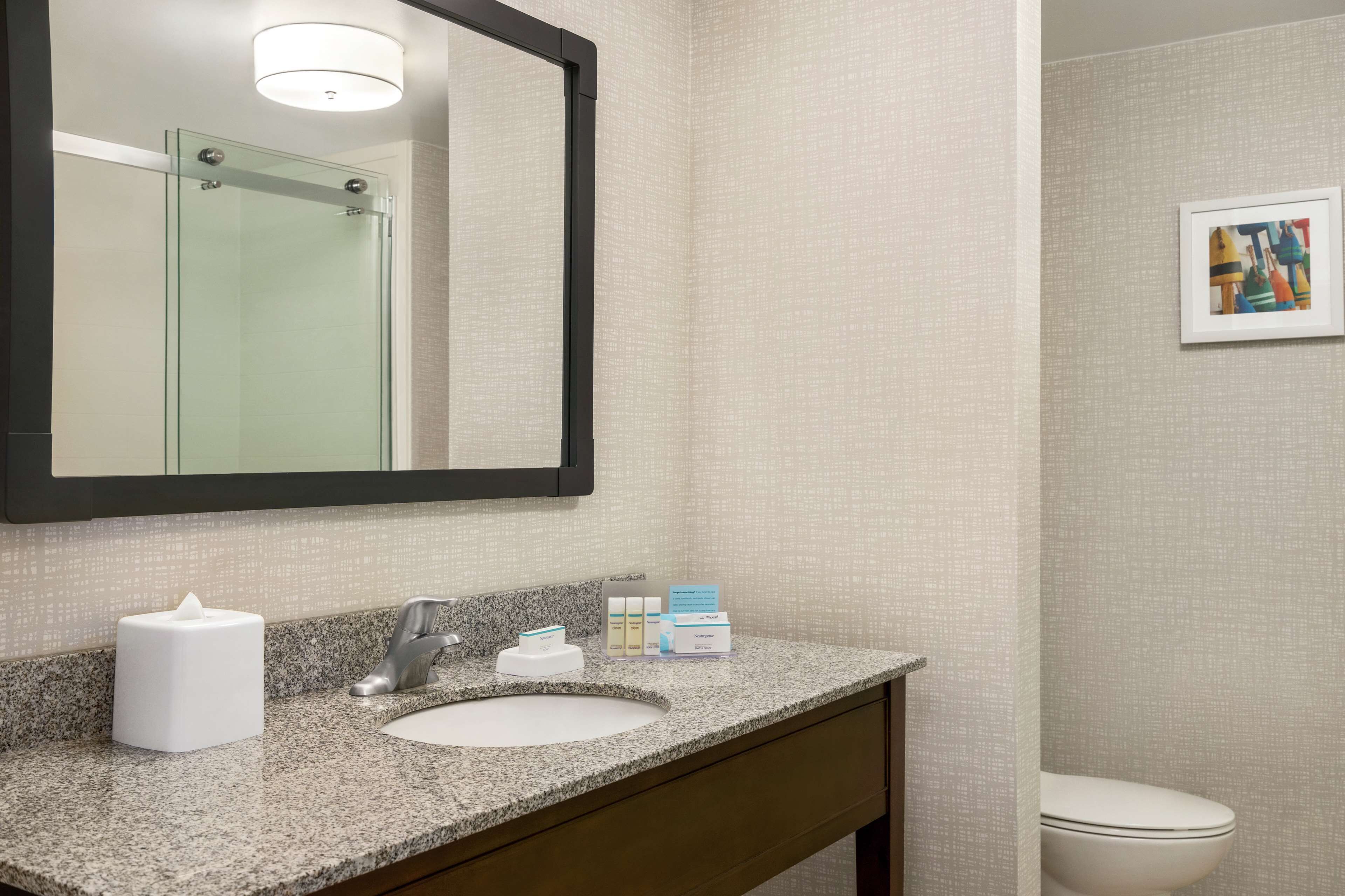 A bathroom with a granite countertop, a white sink, a mirror, and a framed artwork on the wall.