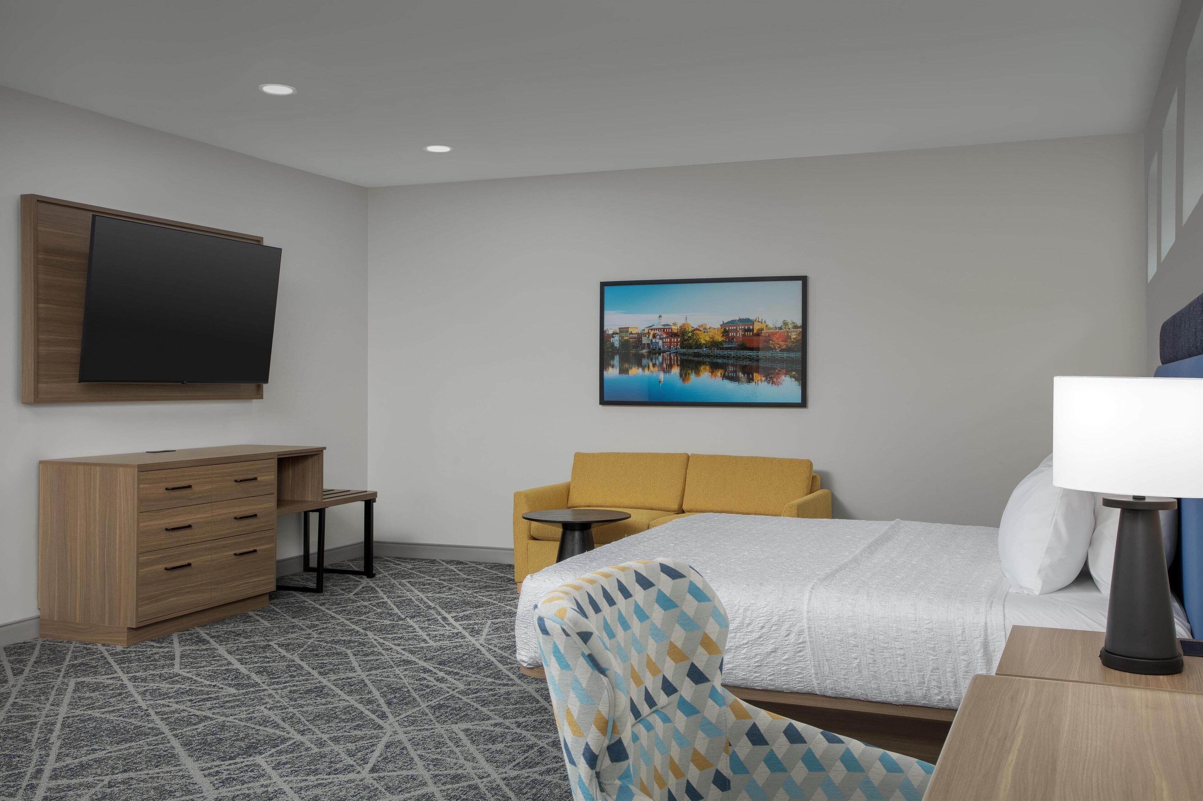 A hotel room with a bed, a sofa, a TV, a desk, and a painting on the wall.
